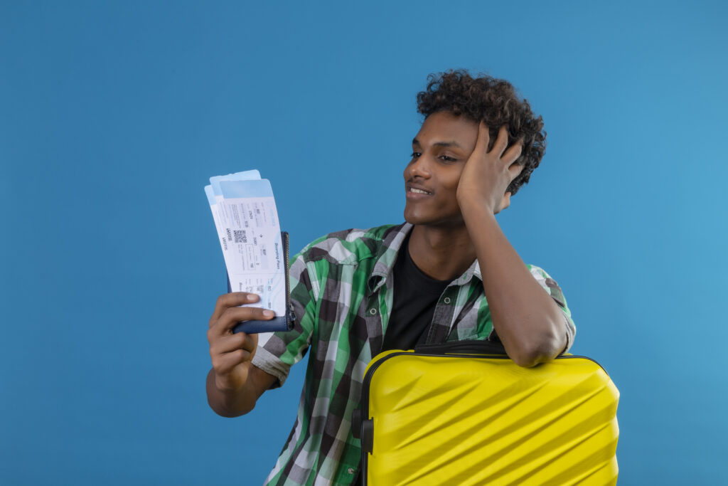 young african american traveler man standing with suitcase holding air tickets looking at them with confident smile on face, satisfied over blue background